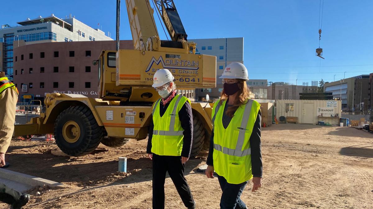 Rep. Melanie Stansbury and a male worker wearing yellow vests and white hard hats walk past a construction crane.