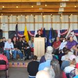 Melanie Stansbury speaks at a podium with American flag bunting as veterans bow their heads behind her.